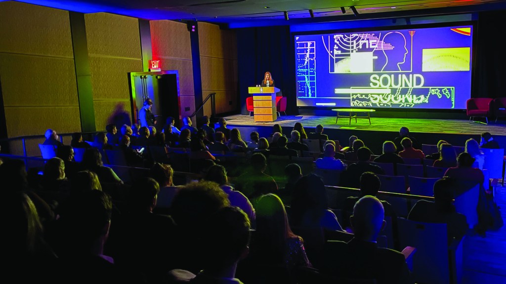 A person on stage addressing an audience in front of a graphic showing intelligence software with the text "SOUND"