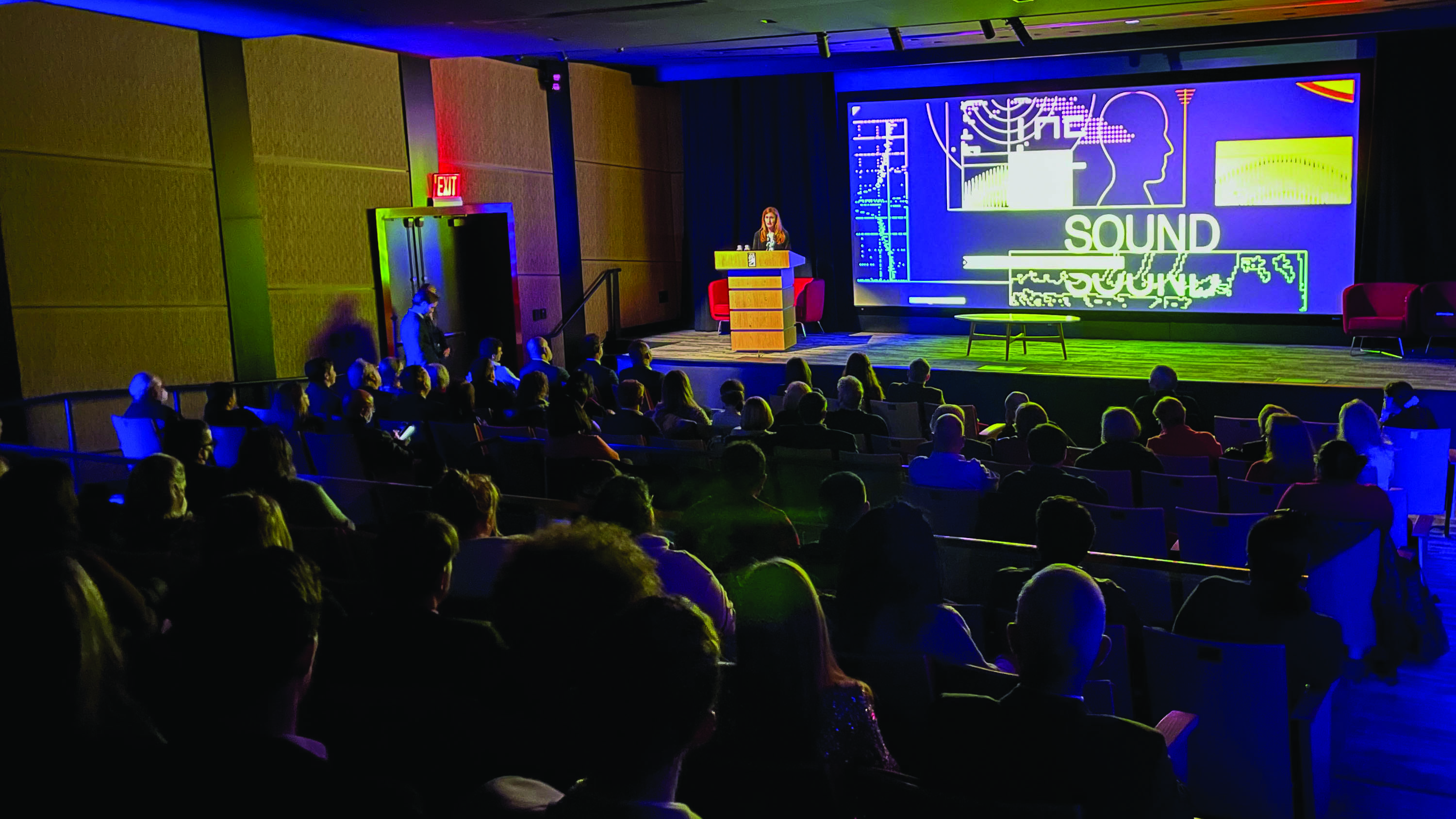 A person on stage addressing an audience in front of a graphic showing intelligence software with the text "SOUND"