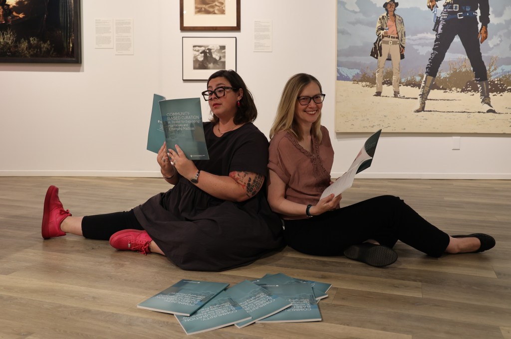 Two  people sitting on the floor of an exhibition room, smiling and reading a printed toolkit publication next to a stack of copies. 