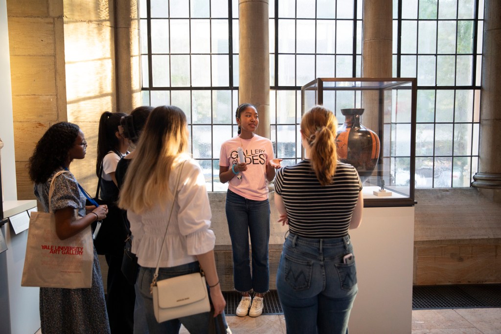 A person wearing a t-shirt that says "Gallery Guides" speaking to a group of people in front of a vase in a vitrine