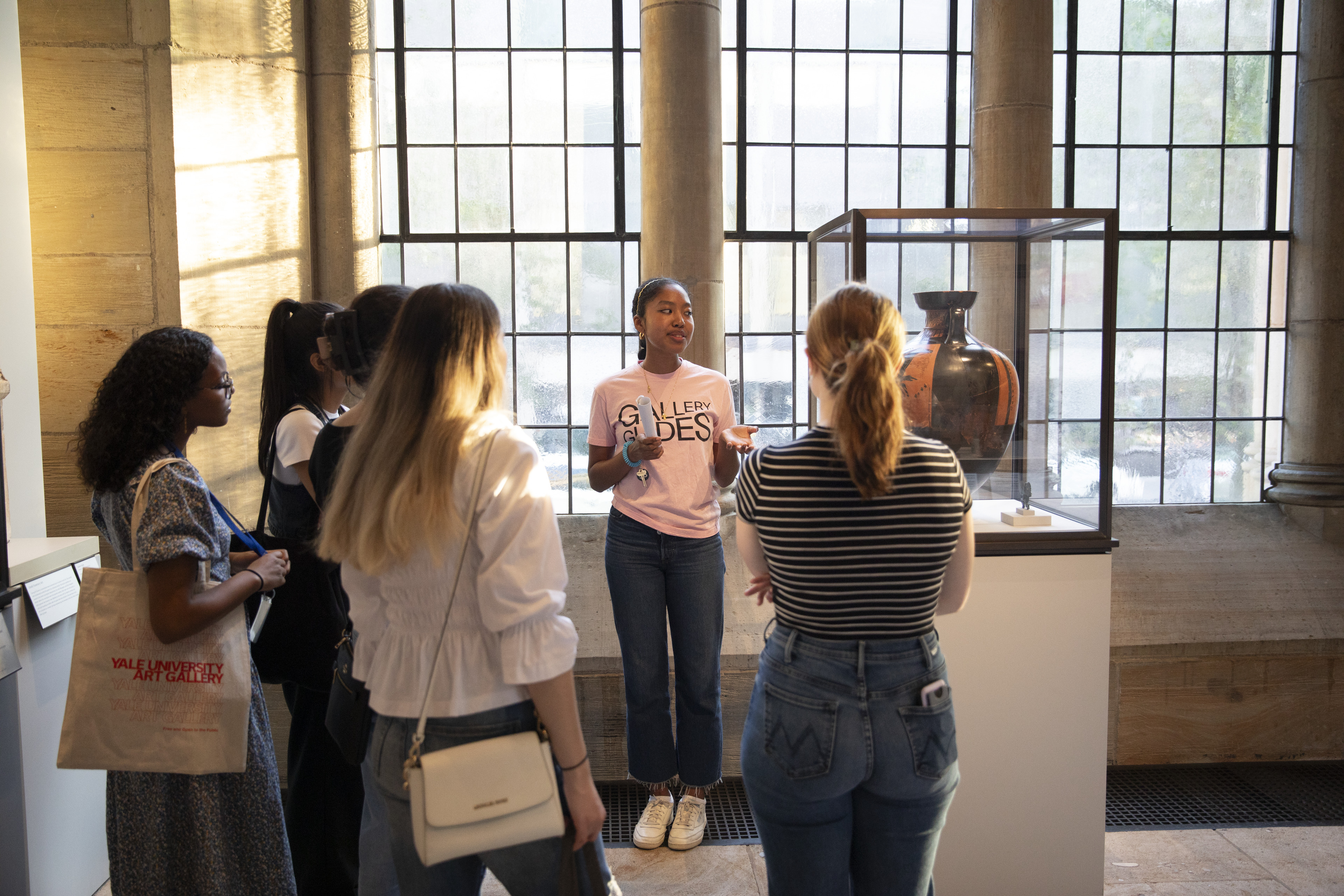 A person wearing a t-shirt that says "Gallery Guides" speaking to a group of people in front of a vase in a vitrine