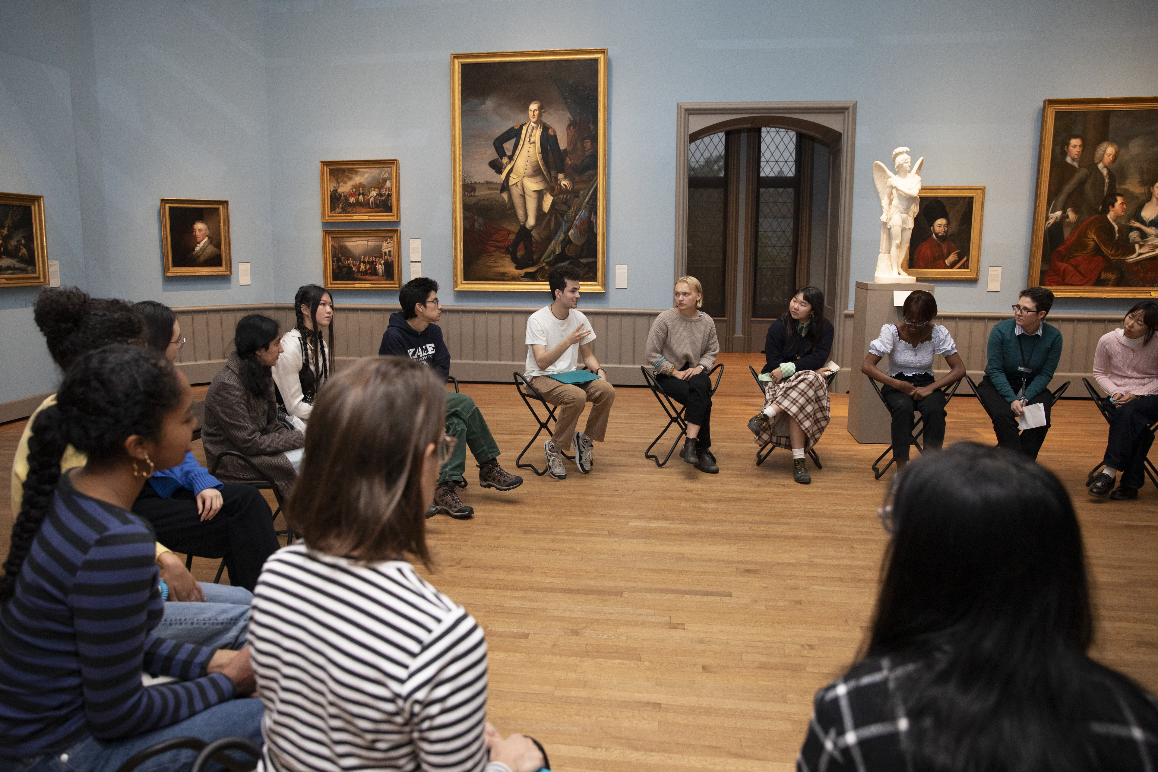A group of people seated in a circle on folding chairs in a museum gallery
