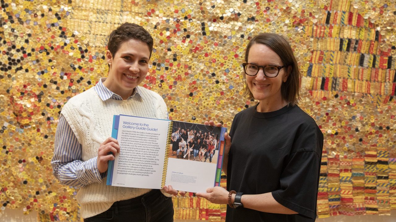Two people in front of a gold mosaic-style artwork holding up a printed publication