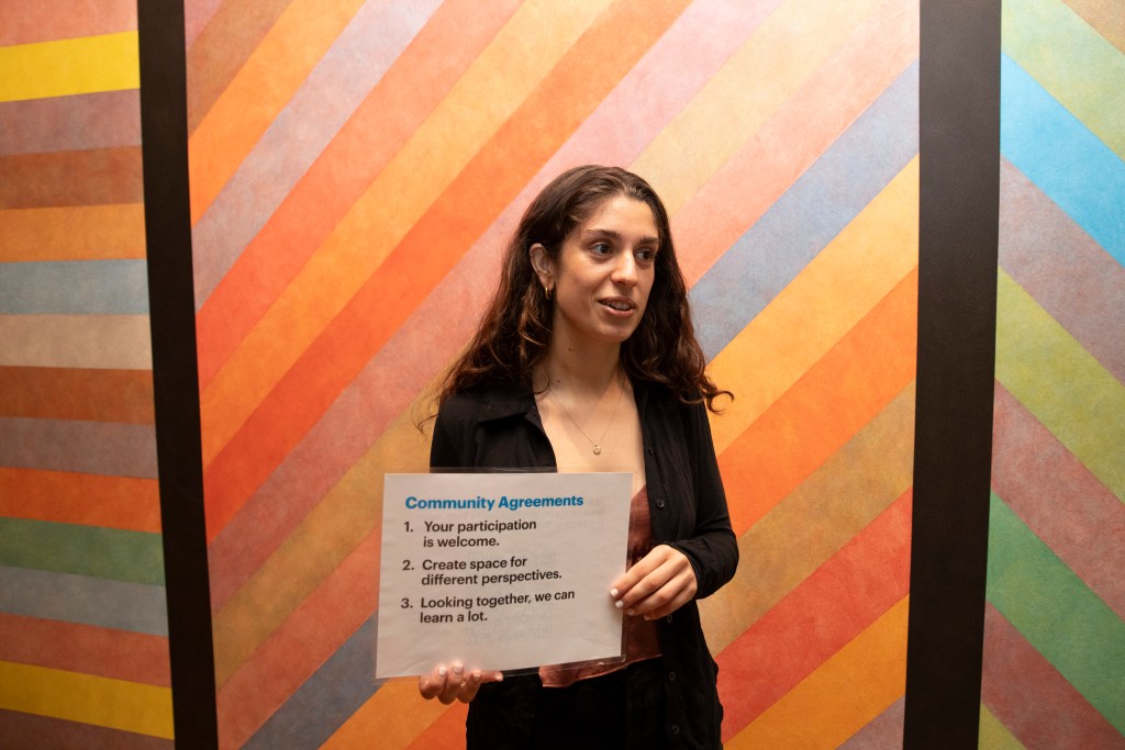A person in front of an artwork holding a sign that reads "Community Agreements: 1. Your participation is welcome. 2. Create space for different perspectives. 3. Looking together, we can learn a lot."