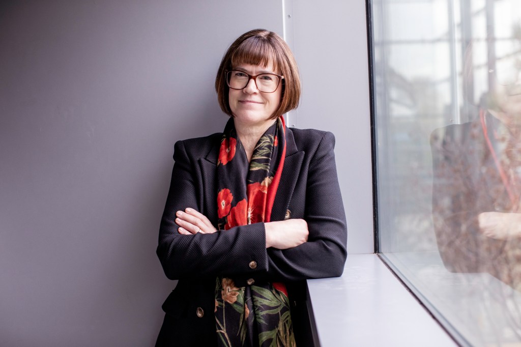 Photo of Lori Bettison-Varga. She is standing next to a window with short brown hair and glasses.