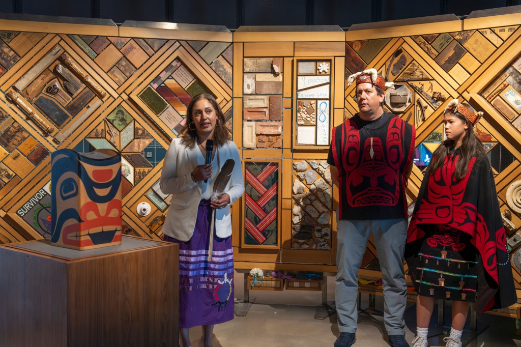 In front of the exhibition that has many patchwork material pieces, a person speaks with a microphone while two others in tribal attire stand nearby.