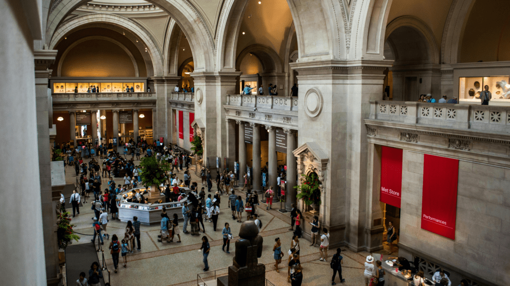 Interior view of the Metropolitan Museum of Art showing a large crowded marble hall with fabric signs for the Met Store and other spaces.