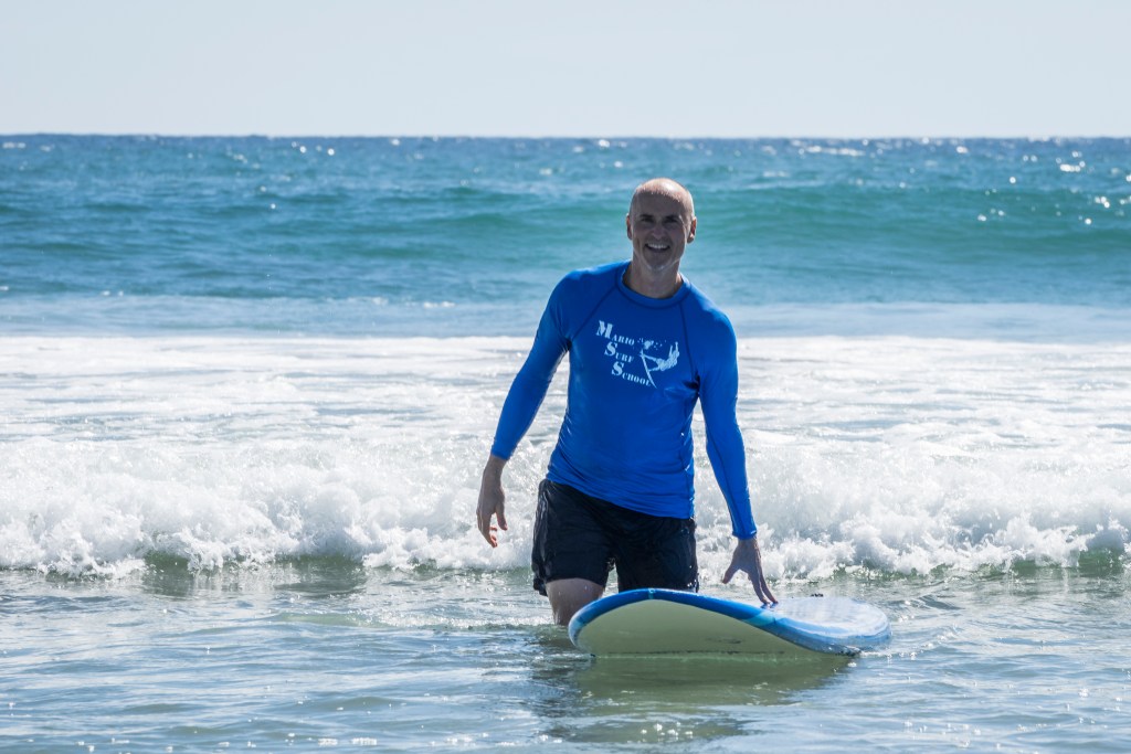 A person standing near the shore of the ocean holding a surfboard and wearing a rash guard reading "Marin Surf School"