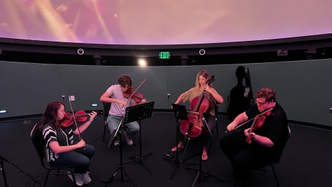 Four musicians in a semi-circle under the glow of a video playing in the planetarium.