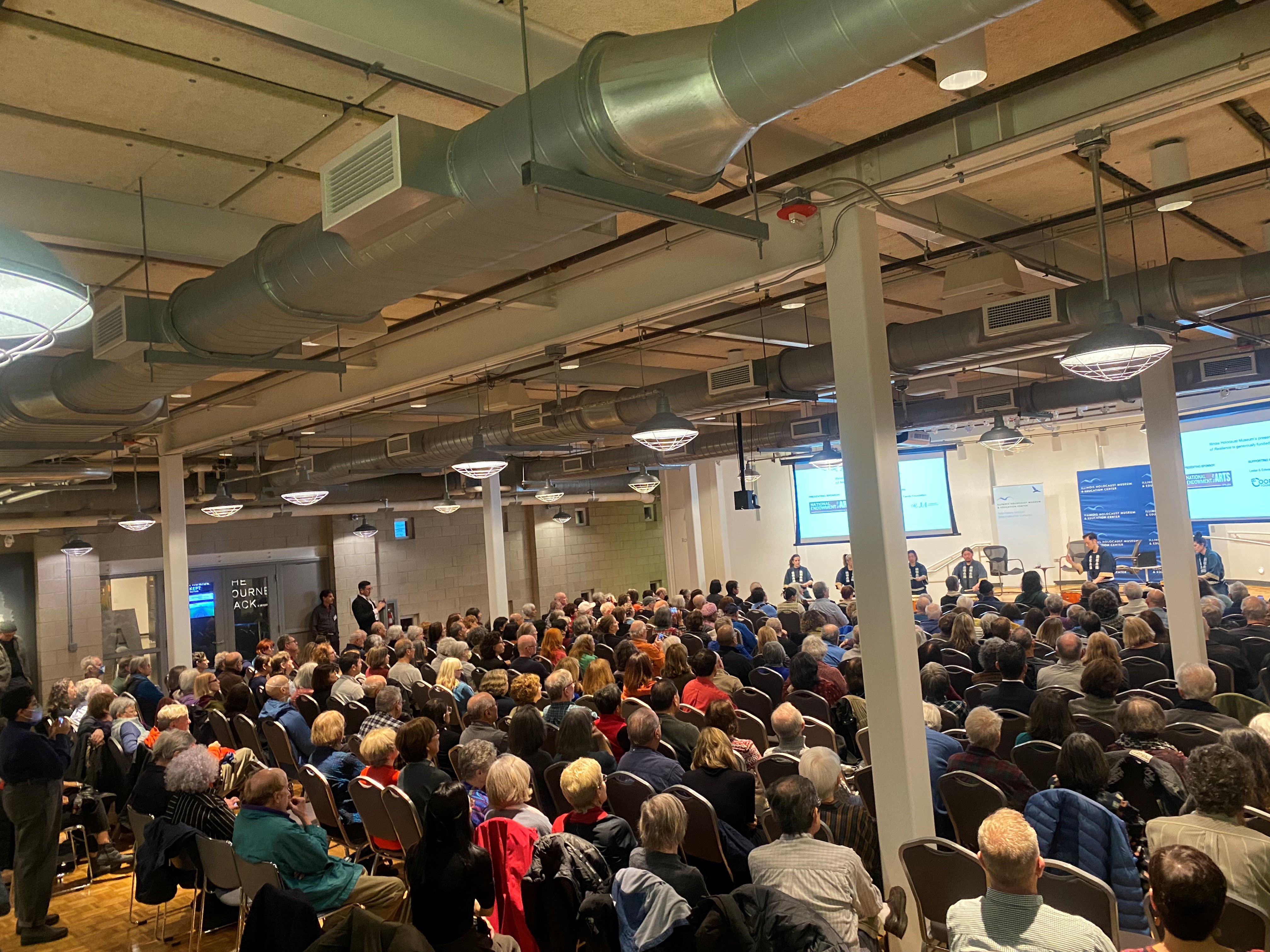 A full crowd of people watching a panel of speakers on stage