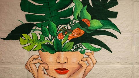 View of a mural on a white brick wall with a female figure that has plants growing out of the top portion of her head.