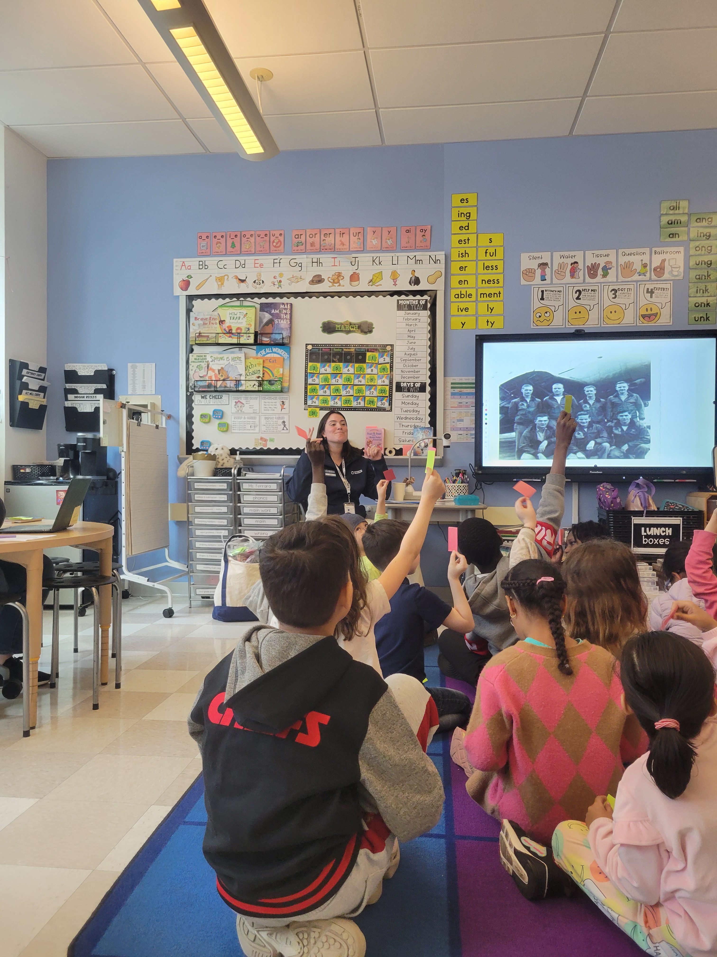 A group of children sitting on a mat in a classroom in front of an instructor showing a black and white group portrait of servicepeople.
