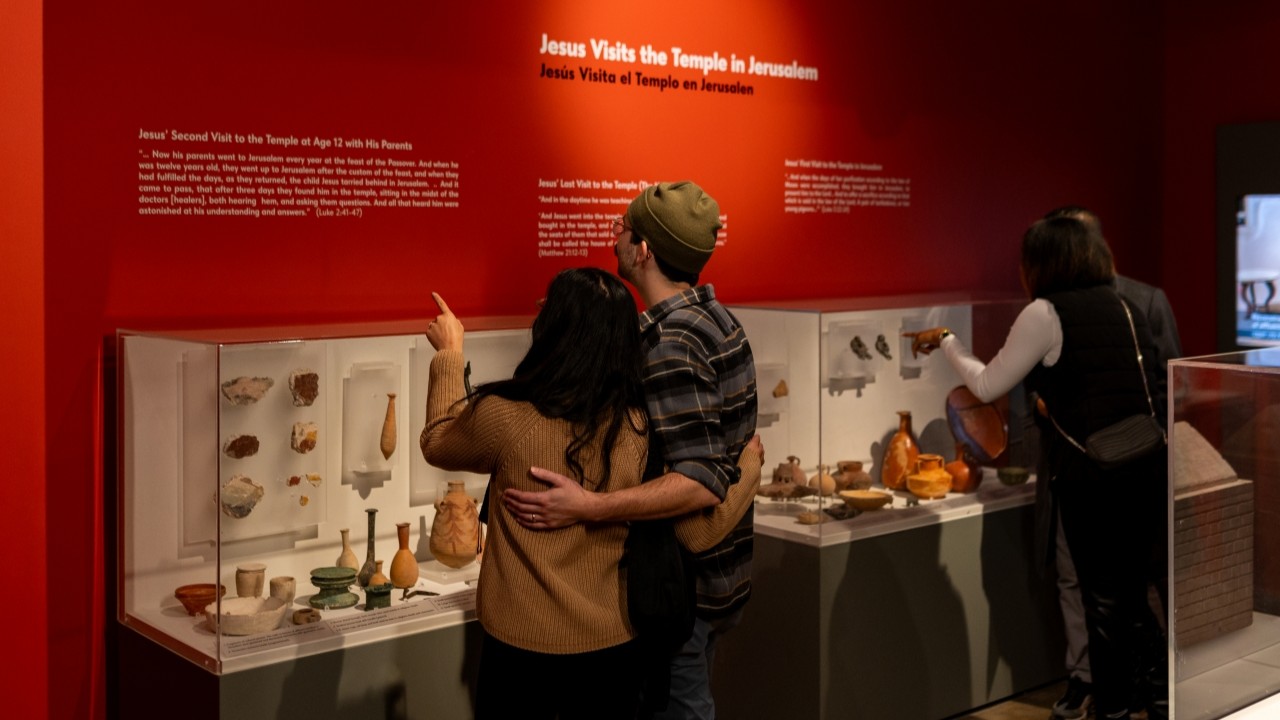 Several visitors look at a text wall with several cases of ancient artifacts displayed. 