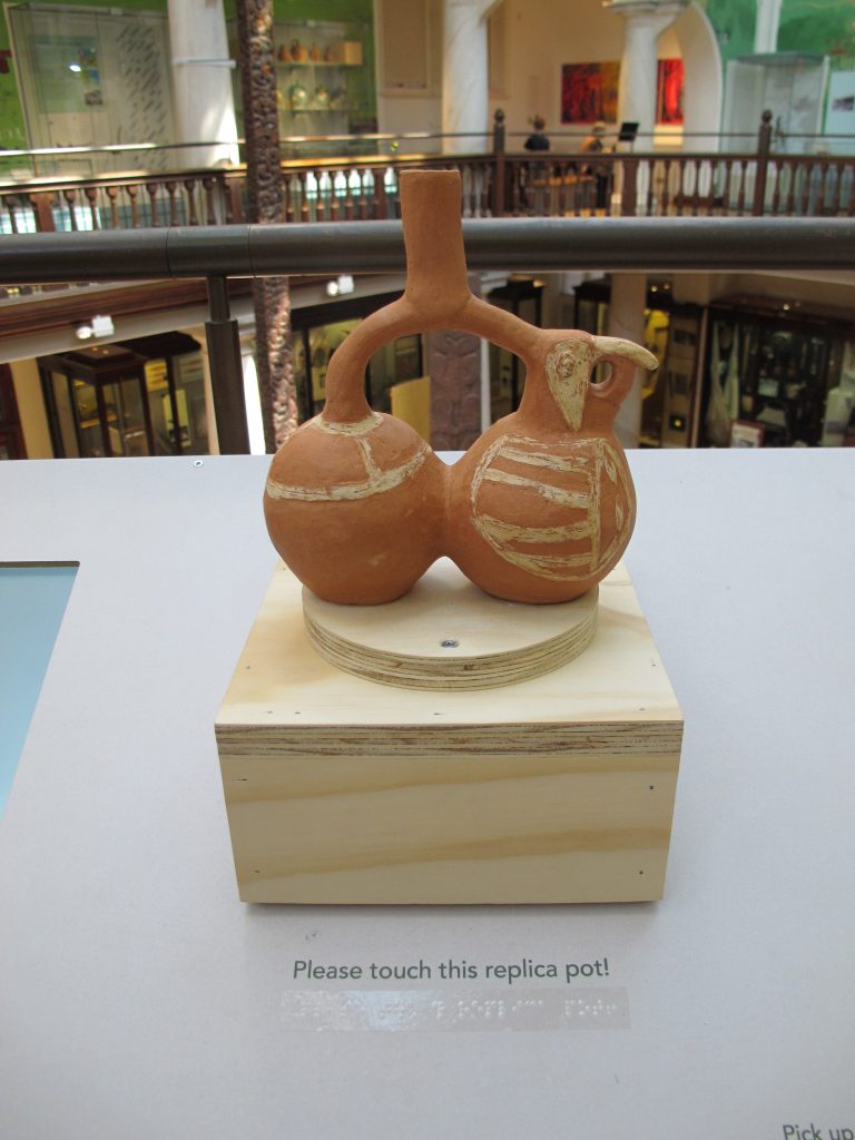 A terracotta replica of a Moche pot sits atop a square plywood base in a museum exhibit. The replica pot is a rich orange-brown color with off-white painted details. A vertical cylinder appears at the top, branching out to connect two interconnected spheres. The sphere on the right has the head of a macaw.