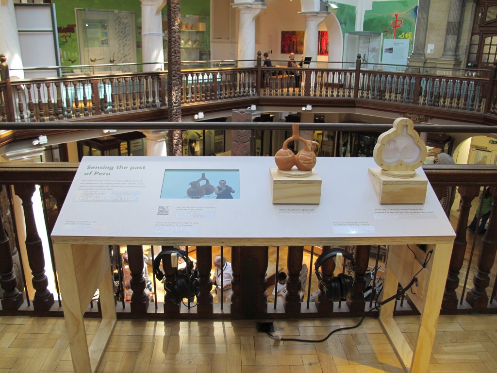 A table-like structure, its top tilted toward the viewer, stands in front of a railing in an ornate museum interior. On the table, there is writing in English and braille, an embedded video screen, and two replicas of a Moche pot. Two pairs of headphones are on hooks at the edge of the structure.