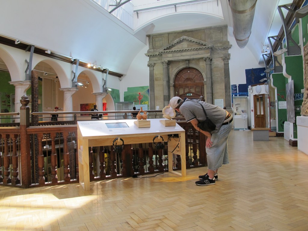 A man in a baseball cap, gray tee-shirt, jeans, and black sneakers stands to the side of a table-top display in a museum. He is bent over to get a closer look at a cross-section tactile of a Moche pot, which he has lifted to move the water inside.