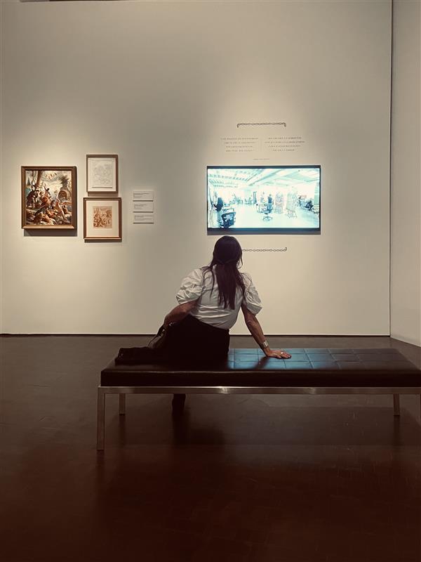 A woman with long, dark hair sits with her back to the camera on a low, armless, backless, leather and metal bench. She leans her weight on her right arm as she watches a video screen on the opposite wall that is hung to the right of a grouping of three works of art.