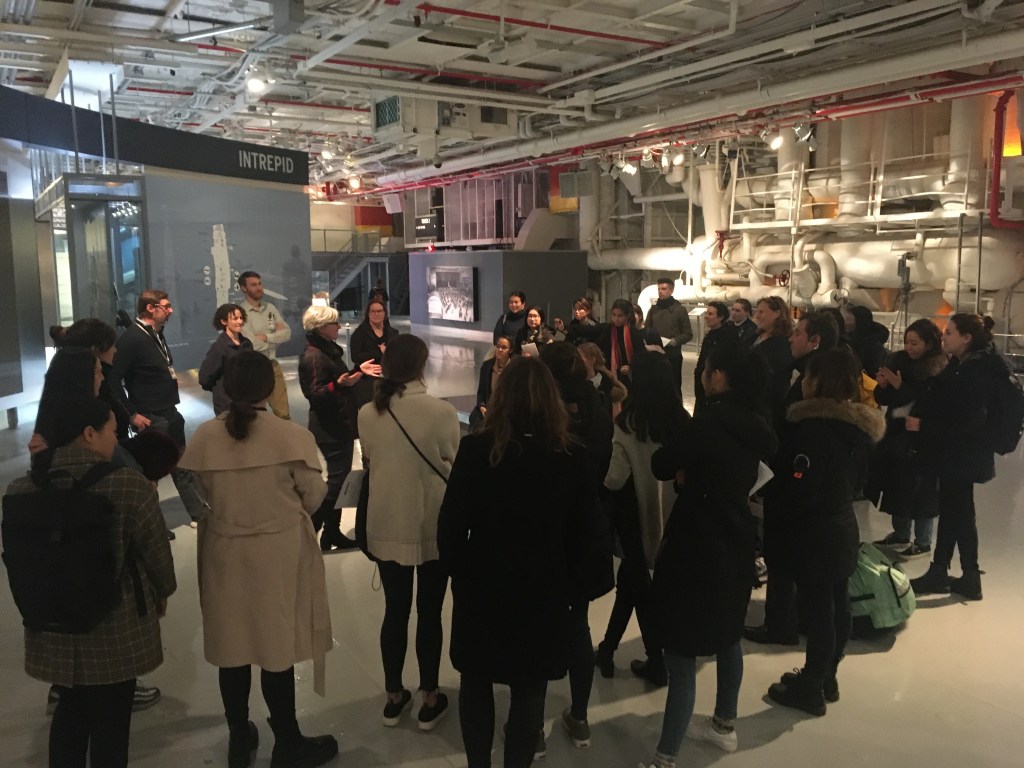 A large group of students gathers inside an open gallery in the Intrepid Museum for their first field trip as part of the NYU course. A woman with short blonde hair stands near the center of the group, addressing them.