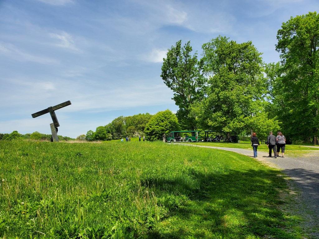 Three figures walk along a gravel path on a beautiful sunny day. A green tram car and additional figures appear farther up the path. To the left, a large abstract steel sculpture resembling a tilted letter T rises from a green field.

