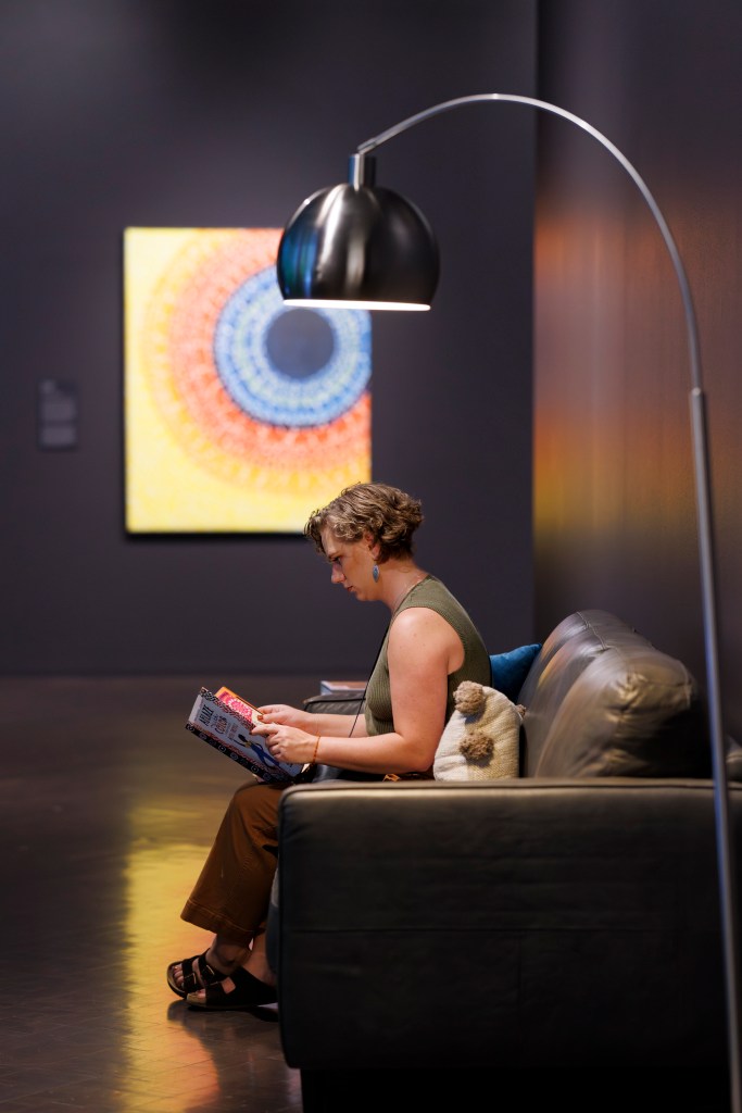 A woman with short, curly brown hair sits on a leather couch with throw pillows reading a children's picture book underneath an arcing metal floor lamp. The camera has captured the woman in profile. On the wall behind her is a large abstract painting. The painting is a vertical rectangle with a dark blue circle in the upper-right portion as its focal point. Colored circles emanate out from the blue center: first white and blue, then orange-red and white, and final yellow and white.