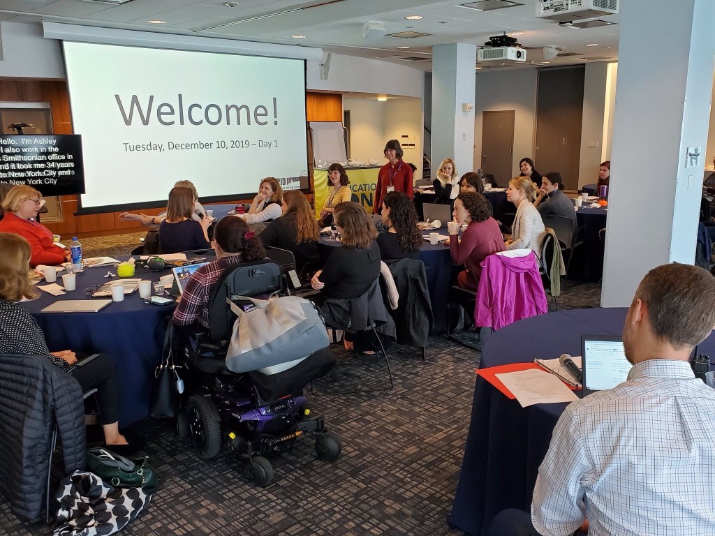 A group of adults, at least one of whom is using a wheelchair, gathers in a brightly lit conference room, seated at multiple tables in small discussion groups. Papers, notebooks, laptops, and food are on the tables, indicating a collaborative workshop or meeting. A screen on the wall displays the word Welcome! and the date, Tuesday, December 10, 2019—Day 1.