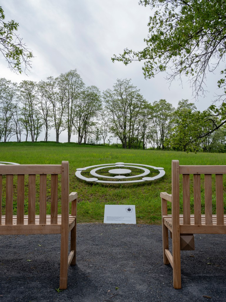 ] An artwork composed of three concentric circles rests low to the ground in a grassy field with tall trees beyond. In front of the artwork, two sturdy wooden benches with backrests and arms have been placed in a leveled, gravel area within viewing distance of the work and its interpretive placard.