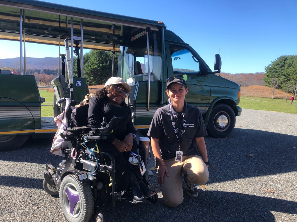 Two women smile for the camera in front of a parked tram car. The woman on the left is using a motorized wheelchair. She has dark skin and hair and is wearing a white baseball cap and large sunglasses. The woman on the left is fair skinned and wearing a navy-blue cap and polo that say Storm King Art Center and khaki pants. She kneels with her staff badge around her neck.  