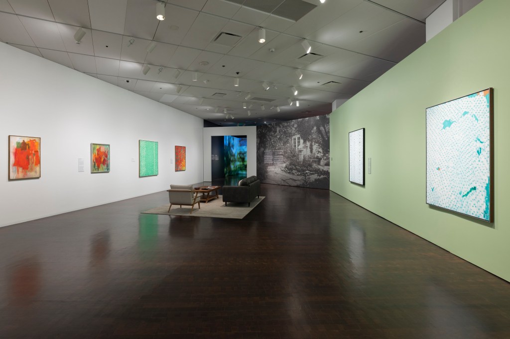 A view into the "Earth and Seasons" section of the exhibition. Thomas's abstract paintings are hung on a white wall to the left and a light green wall to the right. There is a seating area with an area rug, cushioned armchair, coffee table, and leather sofa in the center of the gallery. The sofa is oriented toward the white wall. On the far wall, a floor-to-ceiling black-and-white photographic mural depicts a garden with leafy trees, shrubs, and a paved courtyard. A passageway to the left of the mural leads to another room where blue  and green projections are visible on the walls.