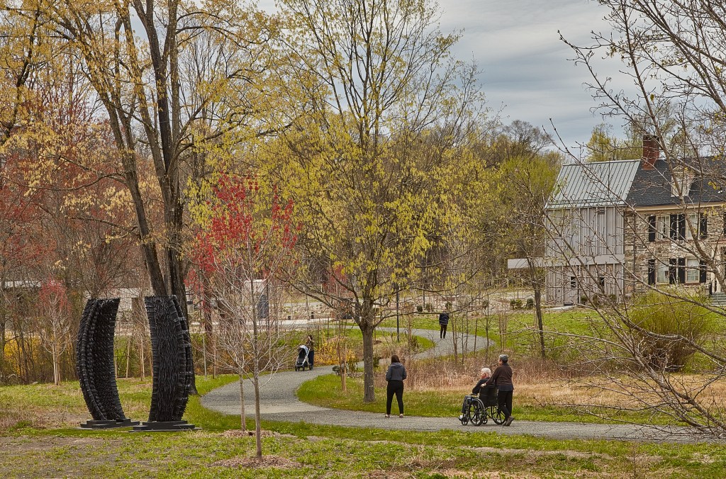 Figures stroll along a winding gravel pathway in early spring. A large sculpture appears to the left, while a stone and metal building is visible through the foliage to the right.