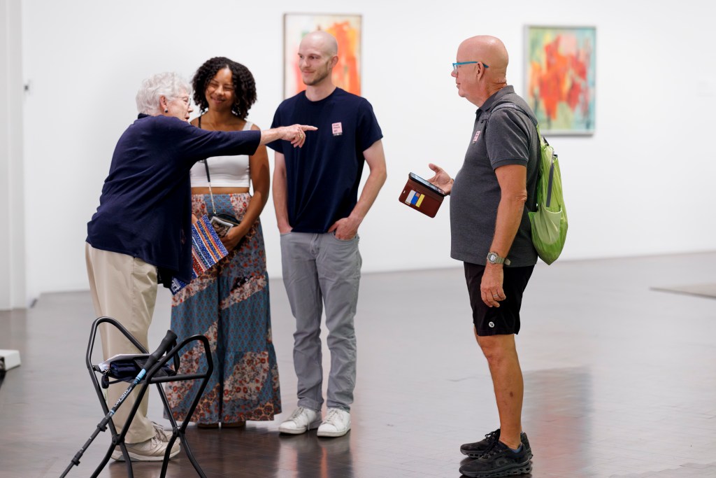 A view into a museum gallery where an older woman with short white hair, glasses, a navy-blue shirt, and khaki pants is speaking with three museum visitors. A young woman with dark hair wearing a cropped white tank-top and flowing patchwork maxi skirt smiles at the woman. She is standing next to a young, bald man with facial stubble in a black tee-shirt, jeans, and white sneakers. It appears from their body language that they are visiting the museum together. An older man stands slightly separate to the right. He is also bald and is wearing bright blue glasses, a dark polo shirt, shorts, and sneakers. He holds his smartphone in his right hand and has a bright green nylon bag on his back. All three guests appear to be listening to the older woman as she points beyond them to something out of the picture's frame to the right. Two of Alma Thomas's abstract paintings appear on the gallery wall behind the group, slightly out of focus. In the foreground, there is a black folding gallery stool, on which rests a cane, small purse or bag, and a few sheets of white paper.

