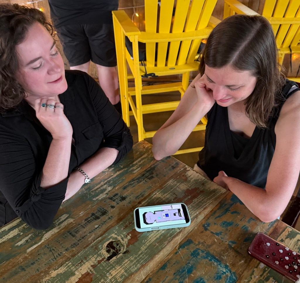 Two women seated at a rustic wooden table, attentively looking at a smartphone lying on the table. The phone displays a 3D rendering of a gallery at the Fralin. Behind them are bright yellow high-back chairs and a person standing at a counter.

