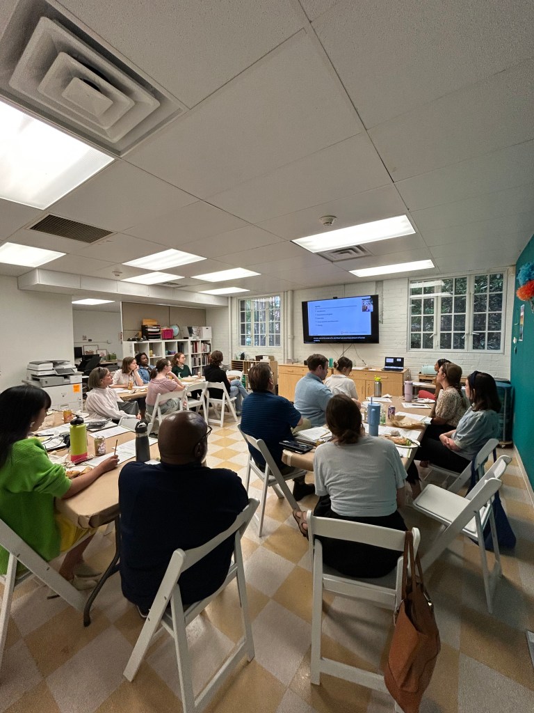 A group of adults gathered in a brightly lit classroom-style space, seated at multiple tables in small discussion groups. Papers, notebooks, and food are on the tables, indicating a collaborative workshop or meeting. A screen on the wall displays a slide with a list, while the teal accent wall in the back is decorated with colorful paper flowers and hanging photographs.
