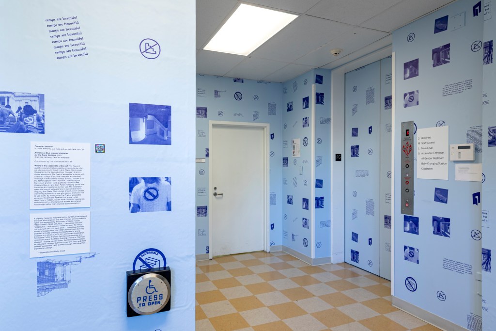 A hallway near an elevator, with light blue wallpaper covered in blue icons, symbols, and text about accessibility. A large round button with the accessibility symbol and the words “Press to Open” is mounted on the wall near the elevator doors. Text panels explain the installation.