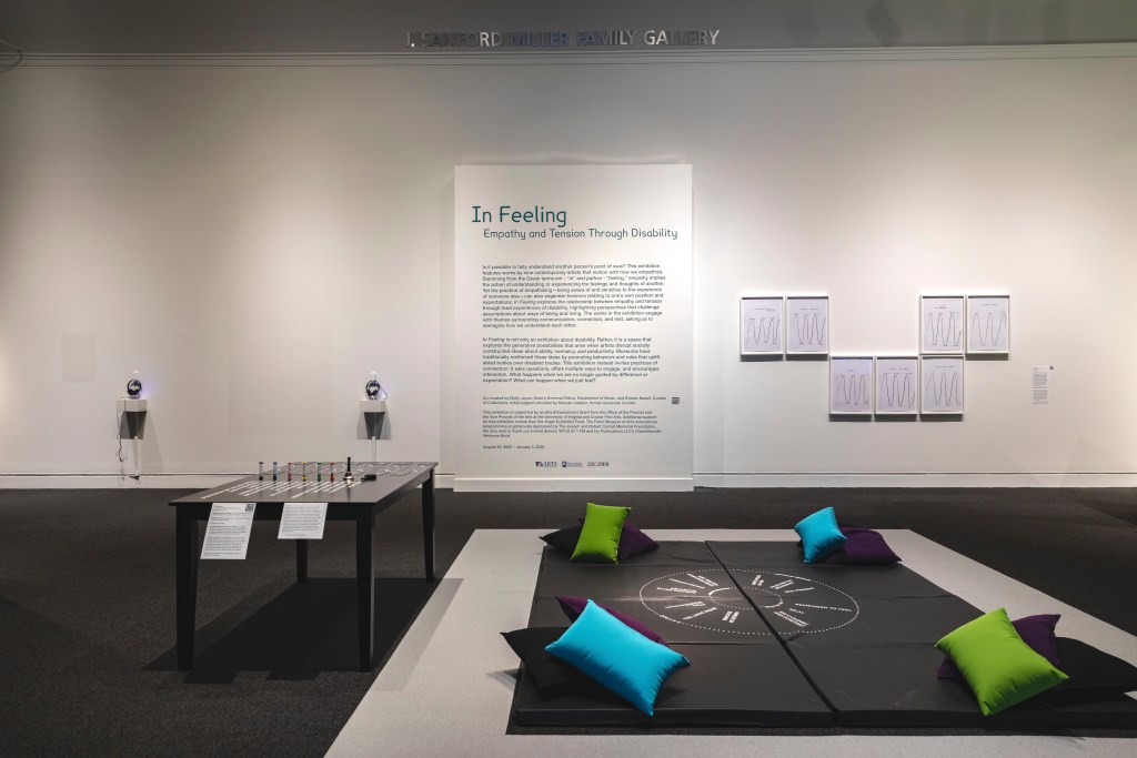 A bright gallery space with an introductory wall text titled In Feeling: Empathy and Tension Through Disability. The installation includes a long black table with interactive elements, colorful pillows on black mats with circles, headphones and subtle white rectangles, and framed line drawings on the wall. The overall setup invites participation and contemplation.