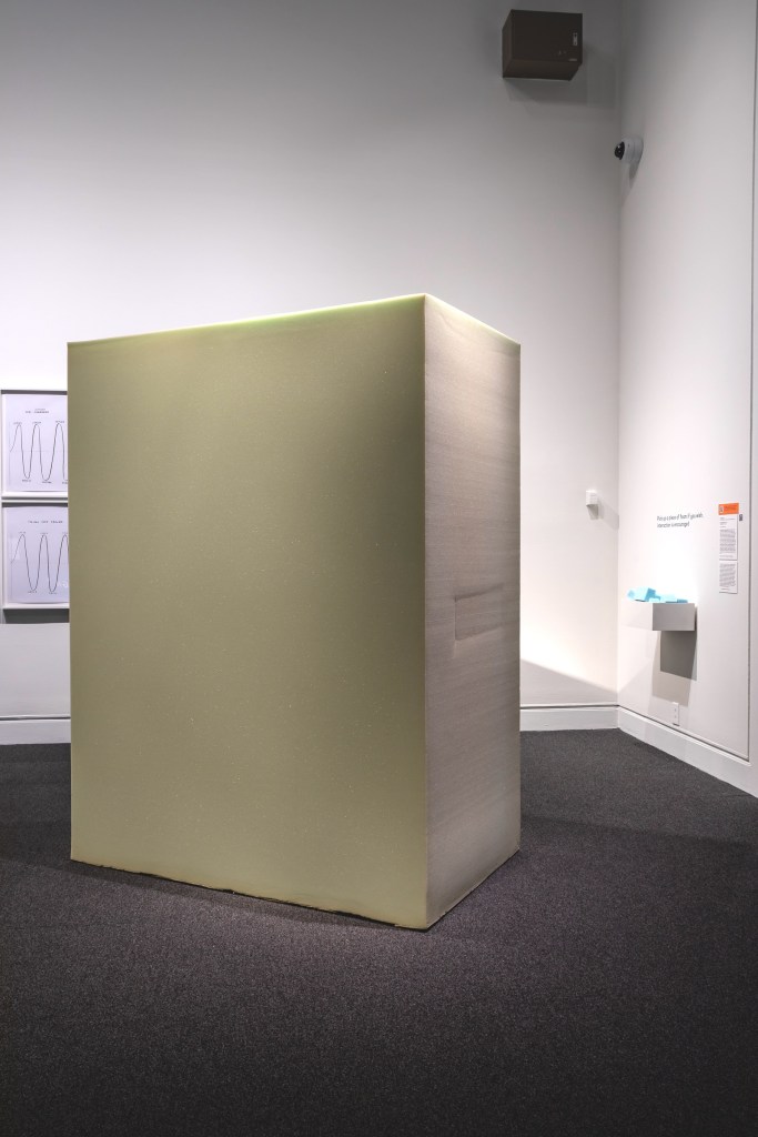 A large, minimalist sculpture in the form of a turquoise block stands in a gallery. Soft lighting highlights its smooth surface, creating a peaceful ambiance. Wall text appears on the right wall, with blue blocks of foam in a box below.

