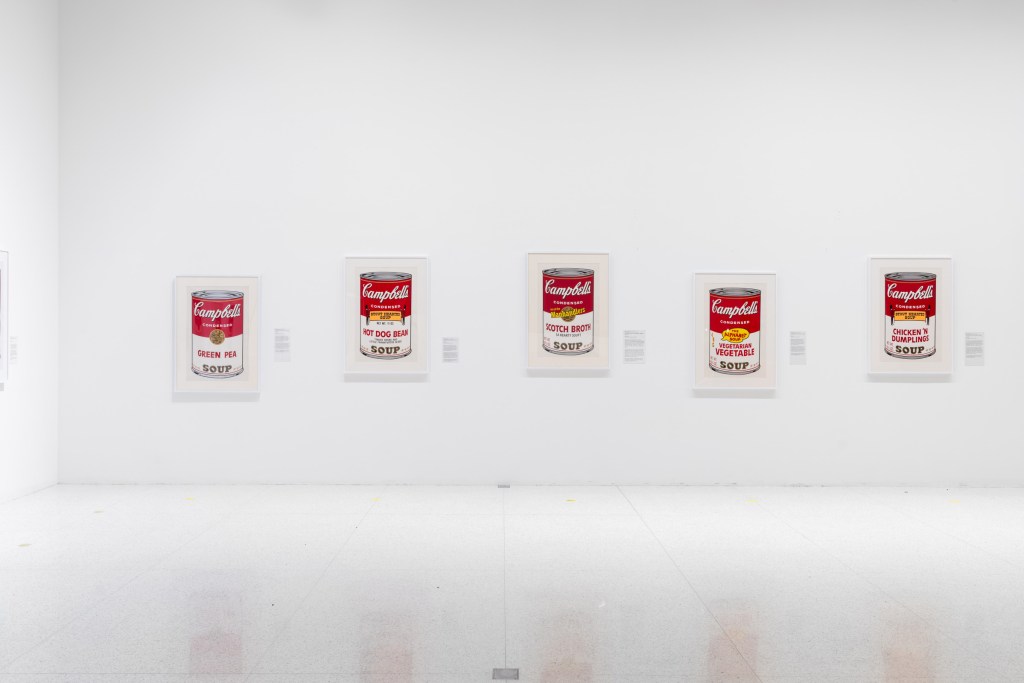Five prints of Campbell's Soup Cans hang on a while wall at varying heights off the floor. Each is accompanied by a wall label hanging to the artwork's right, which are also hung at different heights.


