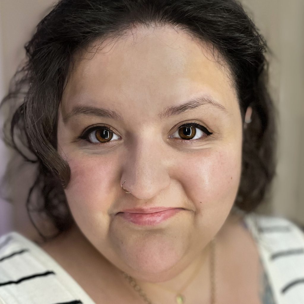 Cropped portrait of an Indian/white British woman with a closed-mouth smile, with short, curly, dark brown hair, brown eyes, and winged black eyeliner. Wearing a pale cream with thin horizontal black striped vest top, two gold necklaces of different lengths, and a tattoo of a bee on the left side of her chest peeking out from the top.