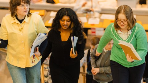 Three teens walk up a flight of stairs holding papers and conversing.