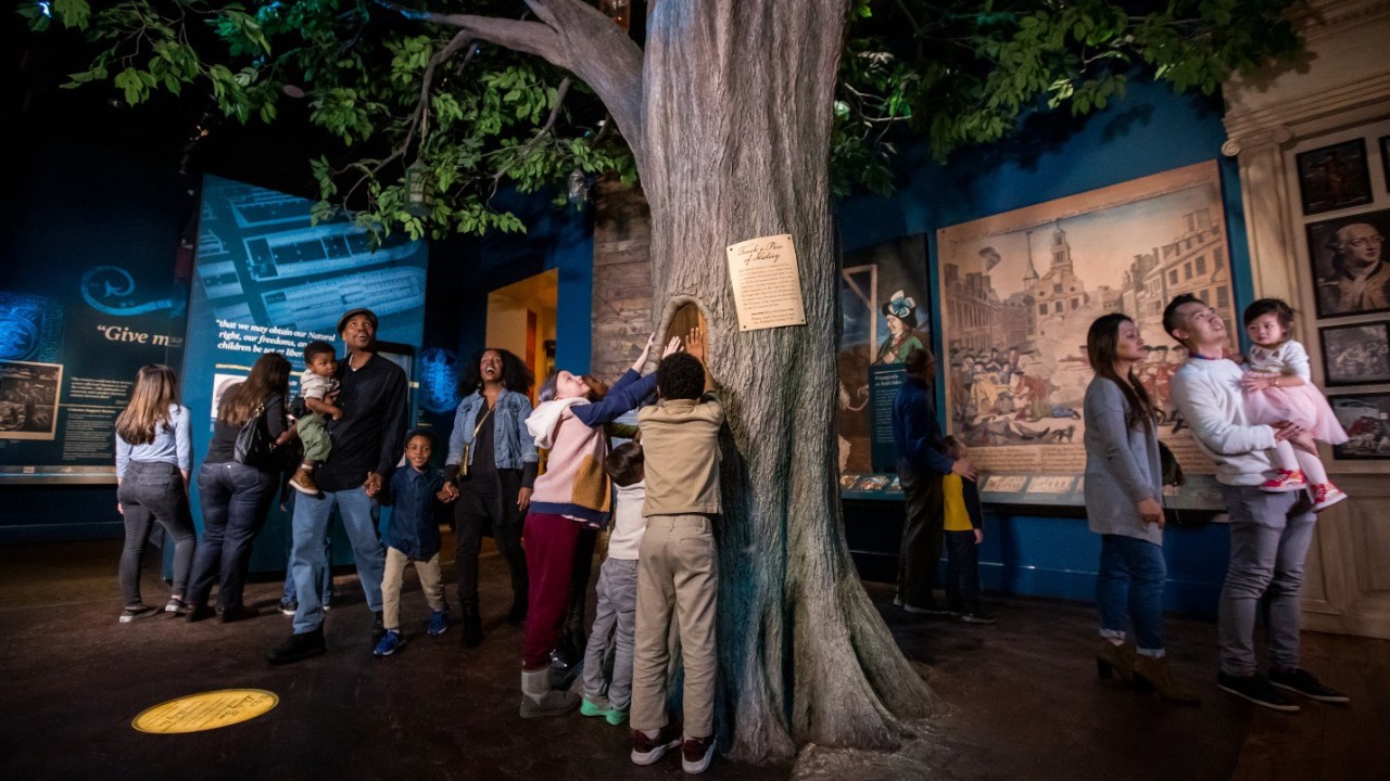 A replica of a tree rises from the center of a crowded museum gallery. Visitors of all ages and appearances talk, walk, and look at the objects and displays around them. A group of children crowd around the tree, touching a sample of real wood that is embedded in the replica's trunk.