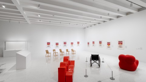 A view into a large gallery with white walls, ceiling, and floor. Ten versions of Campbell's Soup Cans hang a different heights off the floor along the back two walls. Five wooden chairs sit empty in the middle of the gallery. An unoccupied wheelchair sits within stanchions in-between two soft red objects.