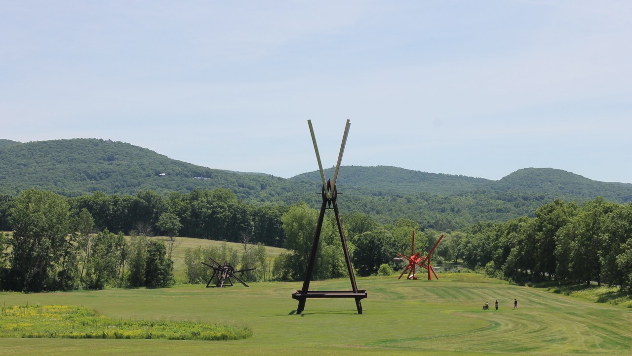 Three monumental abstract sculptures by artist Mark di Suvero stand in a lush green field surrounded by trees and hills. The sculpture closest to the camera is composed of brown and gray steel beams arranged symmetrically to form a triangle with a V on its tip. In the background are two asymmetric sculptures, one brown (left) and one bright red (right). Three people, two standing and one using a wheelchair, are in the field, dwarfed by the size of the sculptures and the vastness of the landscape.