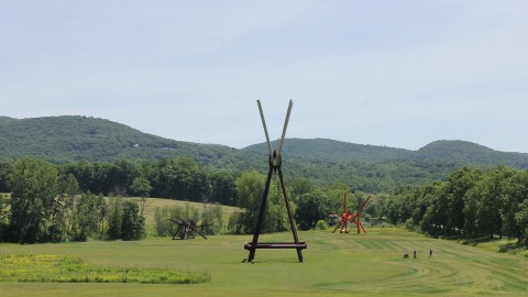 Three monumental abstract sculptures by artist Mark di Suvero stand in a lush green field surrounded by trees and hills. The sculpture closest to the camera is composed of brown and gray steel beams arranged symmetrically to form a triangle with a V on its tip. In the background are two asymmetric sculptures, one brown (left) and one bright red (right). Three people, two standing and one using a wheelchair, are in the field, dwarfed by the size of the sculptures and the vastness of the landscape.