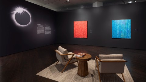 A view into a dramatically lit gallery space with black walls. On the wall to the left, there is a graphic of a solar eclipse to the left of a block of text titled "Space and Sky." On the wall to the right, there are two large abstract paintings. The one to the left is red and white; the one to the right is blue and white with yellow and pink highlights. In the foreground of the gallery, there is a seating area with an area rug, small table, and two comfortable cushioned armchairs. The chairs are oriented so that if a person were to sit in one of them, they would be looking directly at the two paintings on the wall. There are books on the table in-between the chairs.
