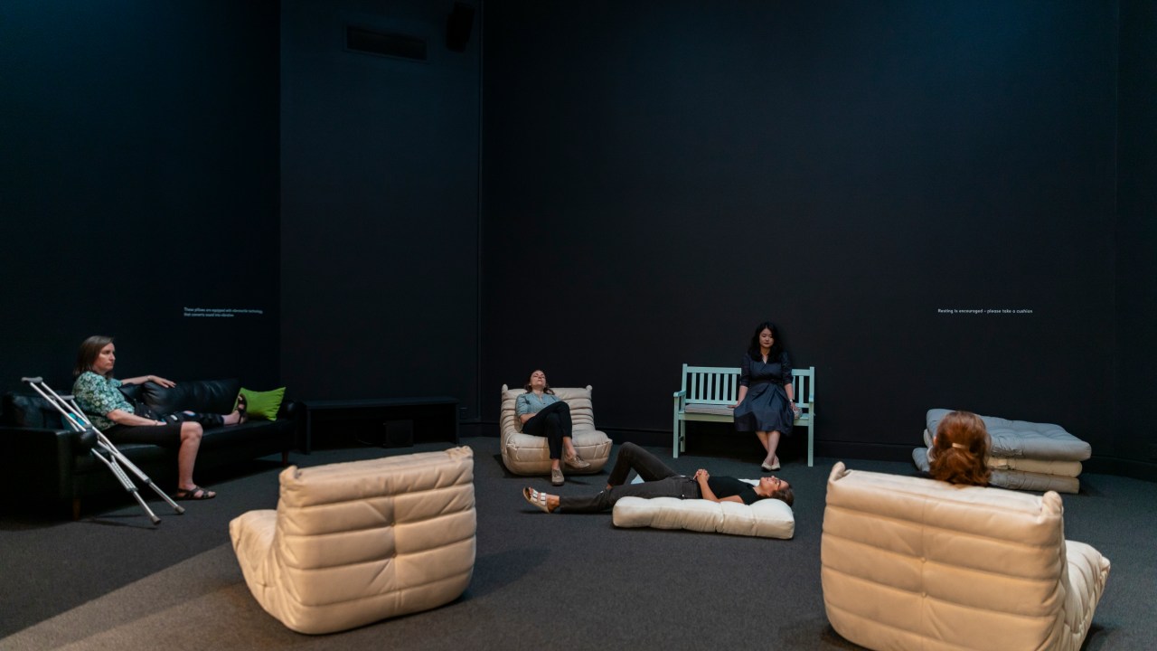 A dark gallery room with scattered, soft cream-colored lounge chairs, a black couch, and a mint-green bench. Five people sit or recline on the furniture, creating a relaxed atmosphere. Short text is visible on the walls, and the space encourages rest and reflection.
