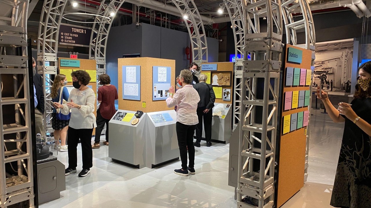 Visitors wearing facial masks interact with a variety of museum exhibits contained within an arching metal scaffold inside the Intrepid Museum. Many of the displays have multisensory components and three visible signs read Content Design Toolkit, Tactile Experiences, and Statistics.