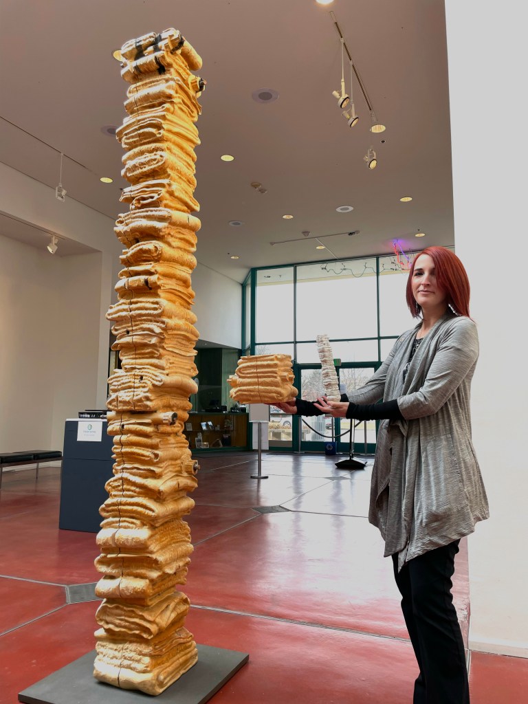 A woman with light skin tone holds two 3D printed tactile objects in her hands next to a tall blonde wooden sculpture of folded blankets in a gallery. The sculpture towers over her, a narrow column of highly textured and carved folded blankets stacked one on top of each other. One tactile object is the same width of the original sculpture but is a short segment. The other tactile objects is a shrunken down replica of the full sculpture and illustrates the height.