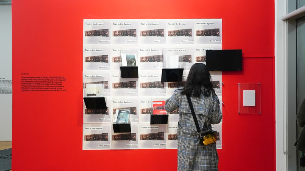 A person stands in front of a large exhibit wall, painted red, with a display of cases with books or magazines, hung on top of a visual background.