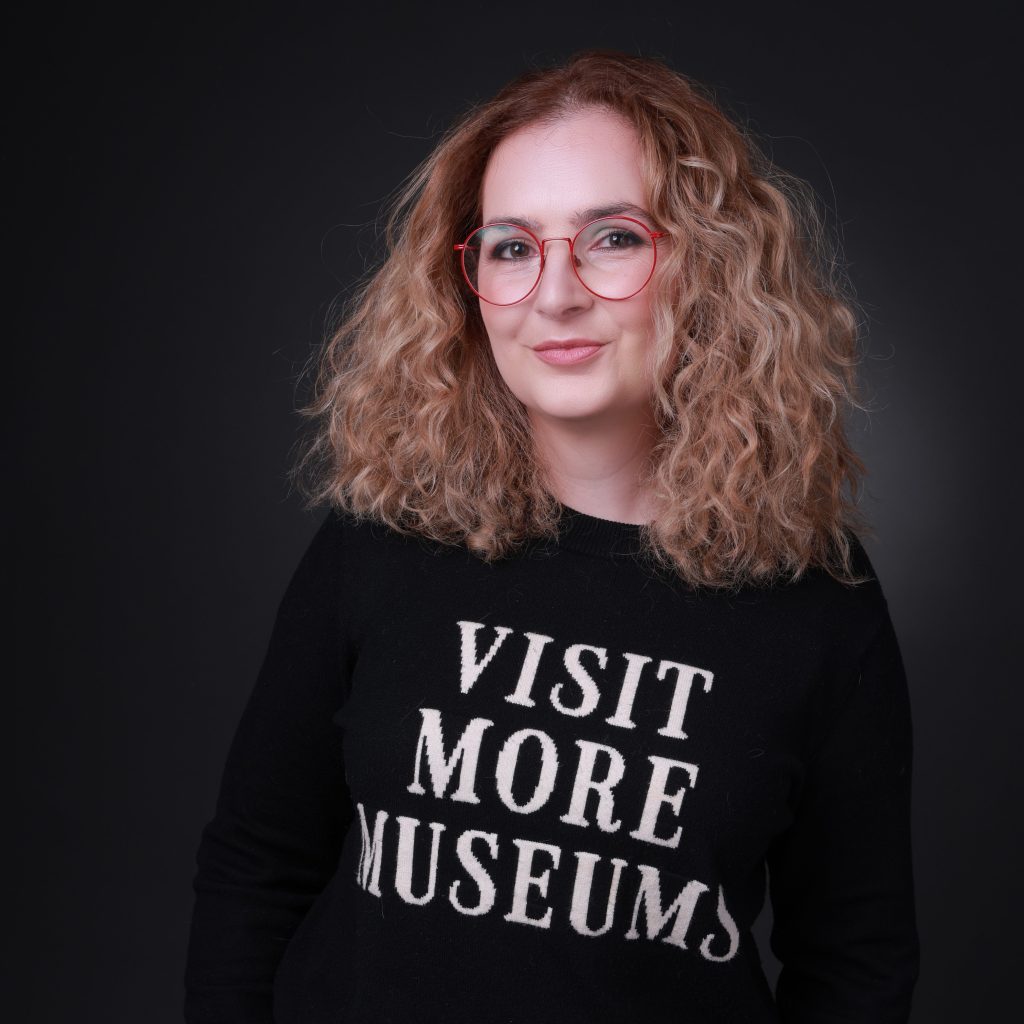 A fair-skinned woman stands against a dark background. She has shoulder-length blonde curly hair and wears red glasses and a black shirt that reads "VISIT MORE MUSEUMS."