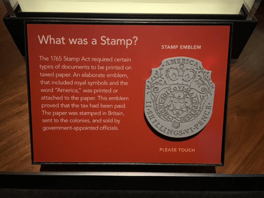 A red museum label with English text and a raised replica of a Revolutionary-era stamp. The label invites visitors to touch the reproduction.

