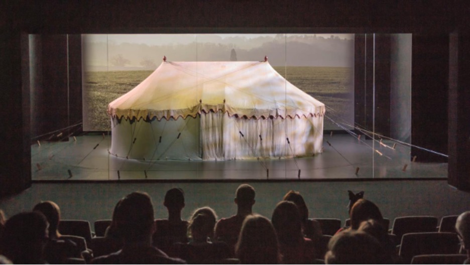 Viewers sit in a darkened theater. An image of a large, oblong-shaped white tent with a peaked roof and many fastening cords appears on screen in front of a landscape backdrop.

